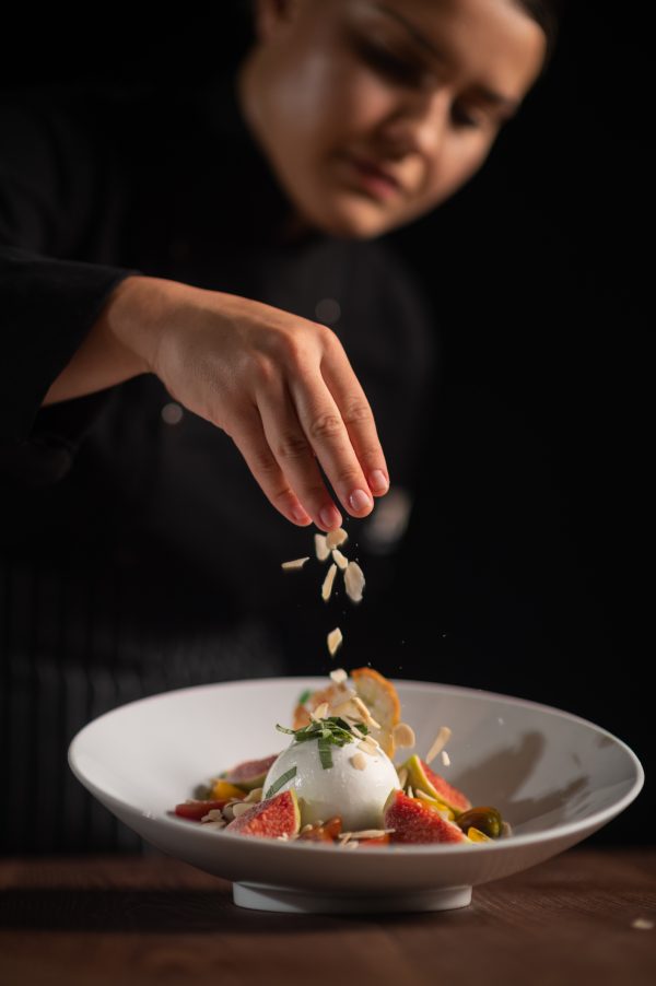 A chef sprinkling sliced almonds over a plated dish topped with burrata and fresh fruit.