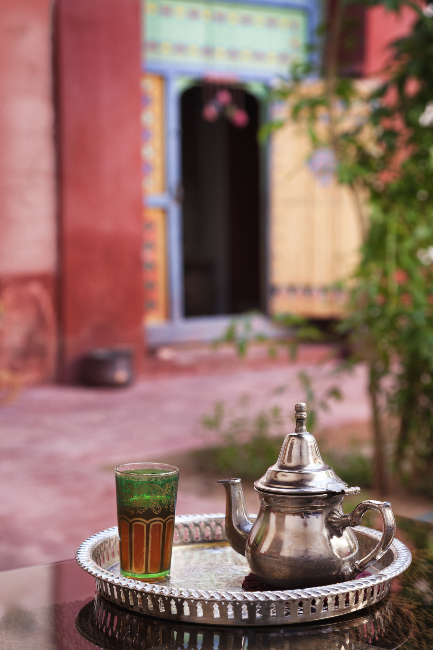 Silver teapot and mint tea in a decorated glass on a metal tray in a Moroccan courtyard with colorful architecture in the background.
