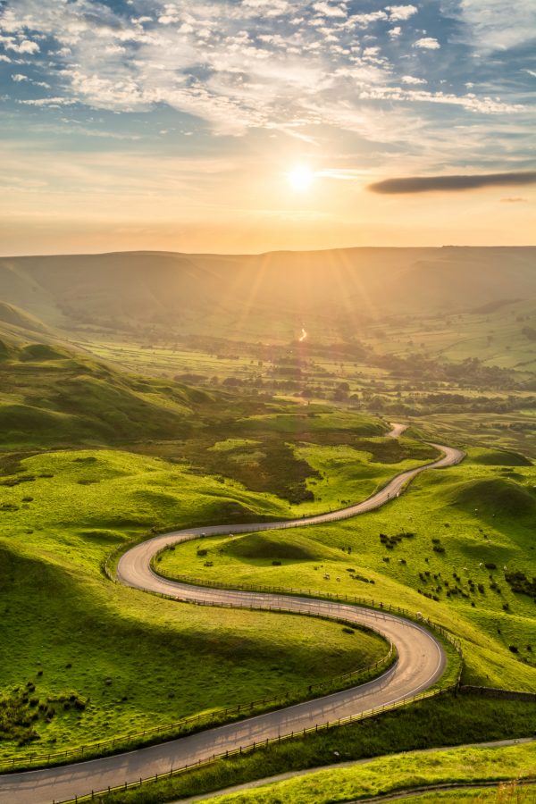 Sunlit winding road cutting through the Peak District under a partly cloudy sky.