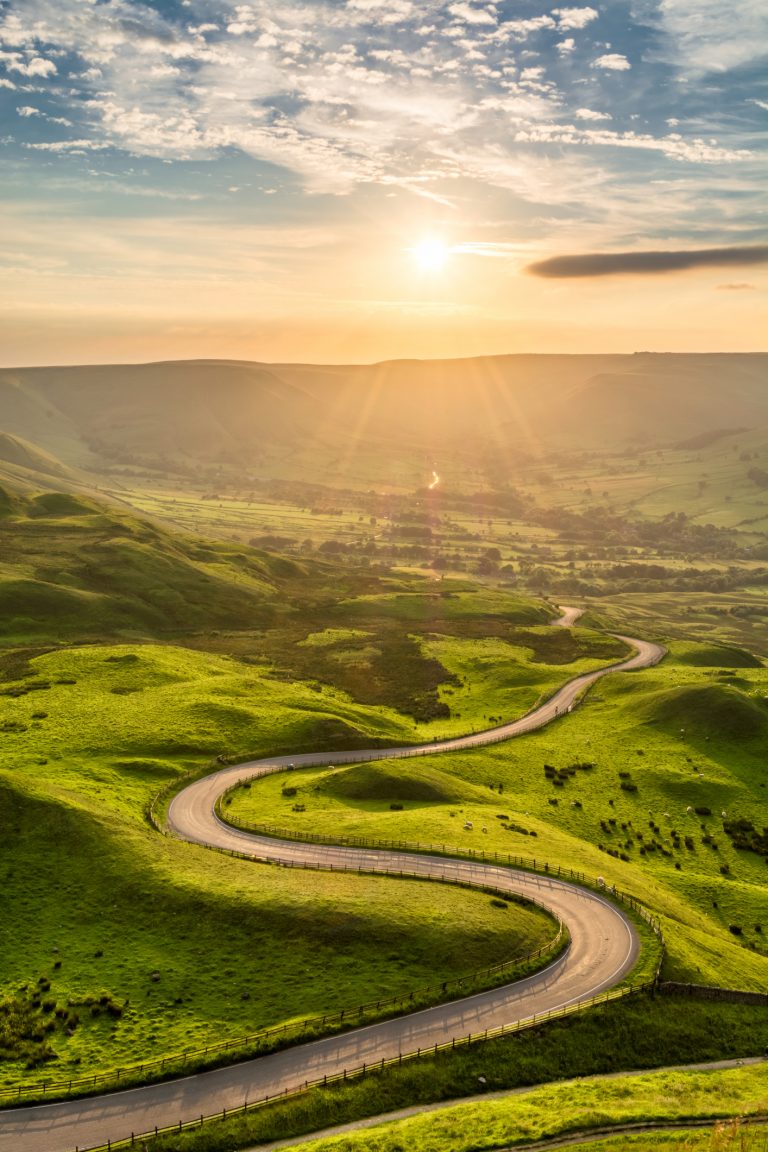 Sunlit winding road cutting through the Peak District under a partly cloudy sky.