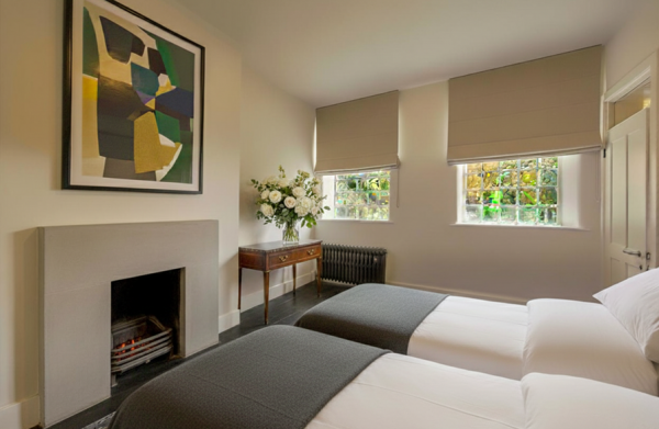 A modern twin-bedded room with a fireplace, abstract artwork, neutral decor, and two windows with roman blinds.