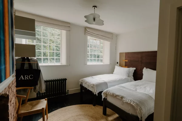 A modern twin-bedded room with a fireplace, abstract artwork, neutral decor, and two windows with roman blinds.