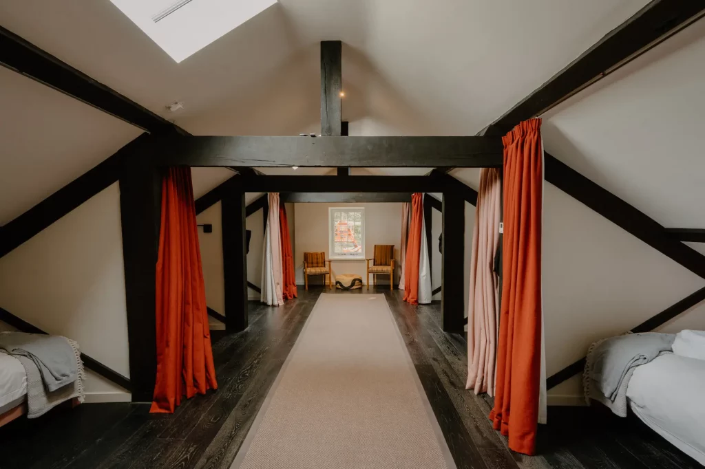 Shared sleeping area with single beds separated by privacy curtains under exposed wooden beams.