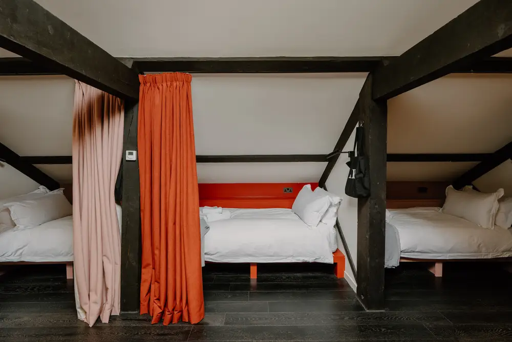 Shared sleeping area with single beds separated by privacy curtains under exposed wooden beams.